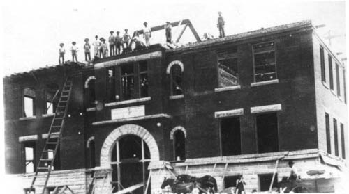 The school, an imposing multi-story brick building, under construction (workmen standing along the roof-line)